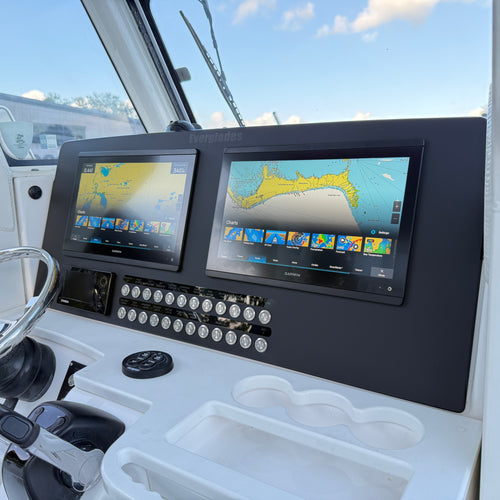 A pair of Garmin GPS navigation screens mounted on a black dashboard inside a boat cabin, with a white console below and outdoor scenery visible through the windshield.